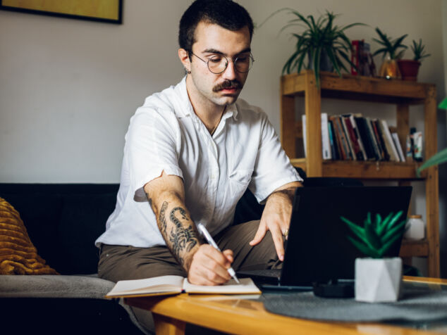Man with a mustache working on laptop at home and writing in an notebook