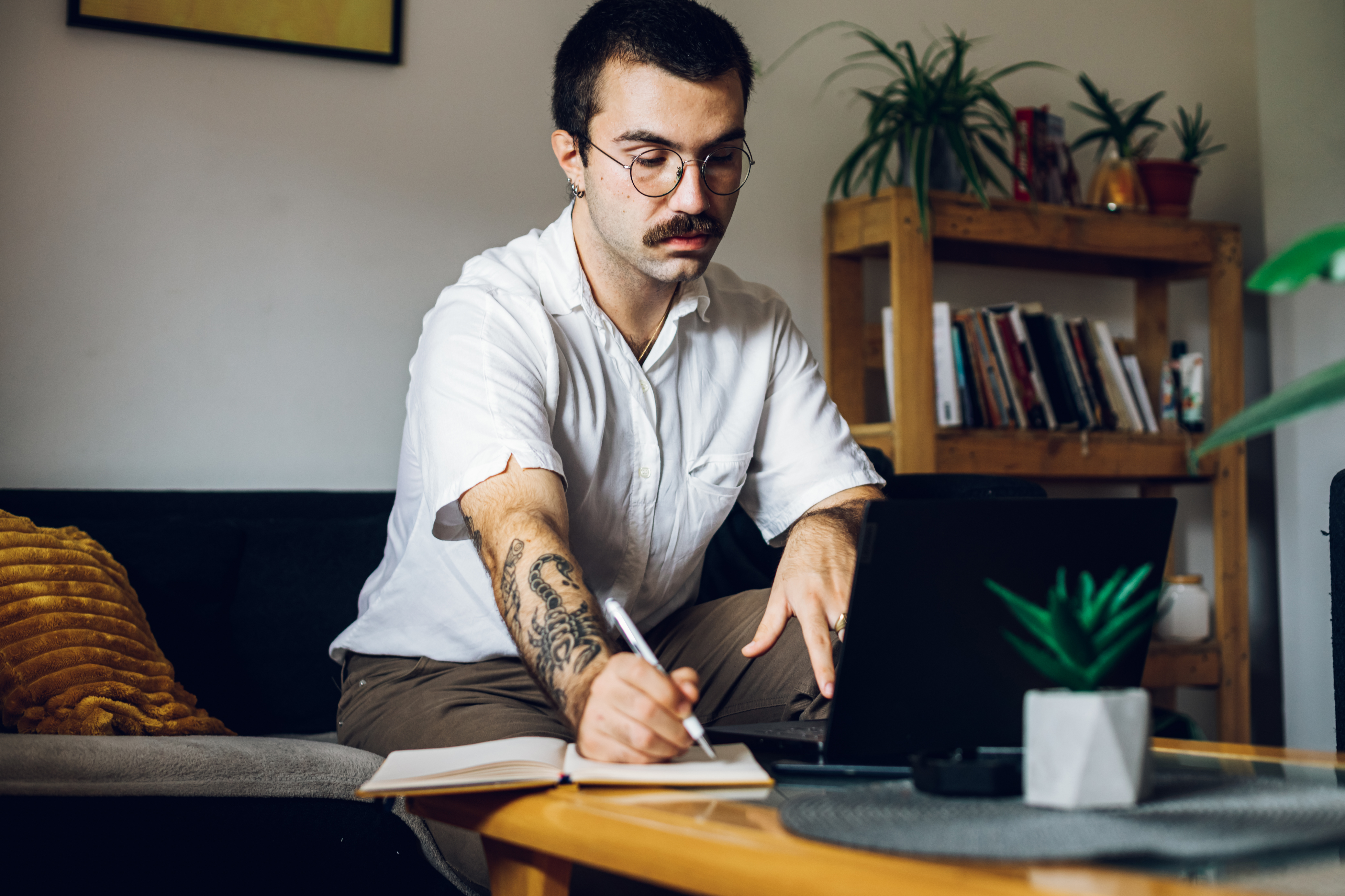 Man with a mustache working on laptop at home and writing in an notebook